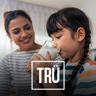 Niña tomando un vaso de leche junto a su madre sonriente con logotipo de TRÜ